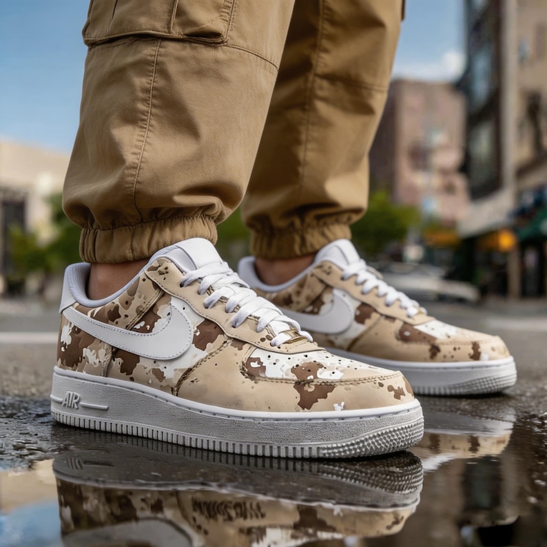 May include: A pair of Air Force 1 sneakers in a desert camouflage pattern. The shoes have a white sole, white laces, and a white Nike swoosh. The sneakers are worn with tan cargo pants.