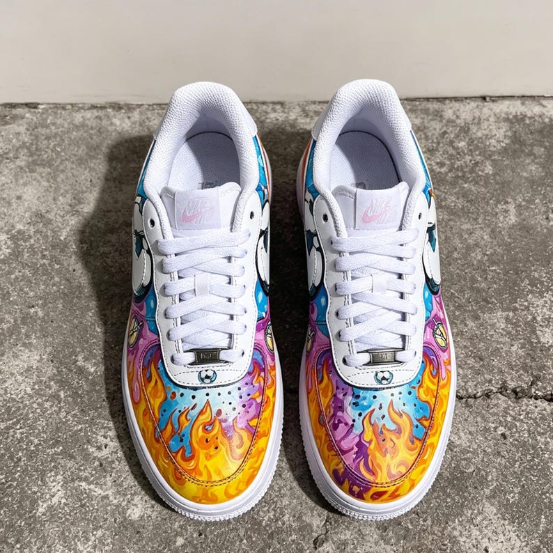 May include: Pair of white sneakers with custom artwork. The design features a colorful flame pattern in yellow, orange, purple, and blue on the toe and sides. The upper part of the shoe has a cartoon design with white laces.