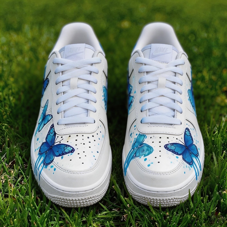 Op de afbeelding: Witte sneakers met blauwe vlinderontwerpen. De schoenen hebben een witte basis met blauwe vlinderillustraties en verfspetters. De veters en zolen zijn wit, tegen een groene grasachtergrond.