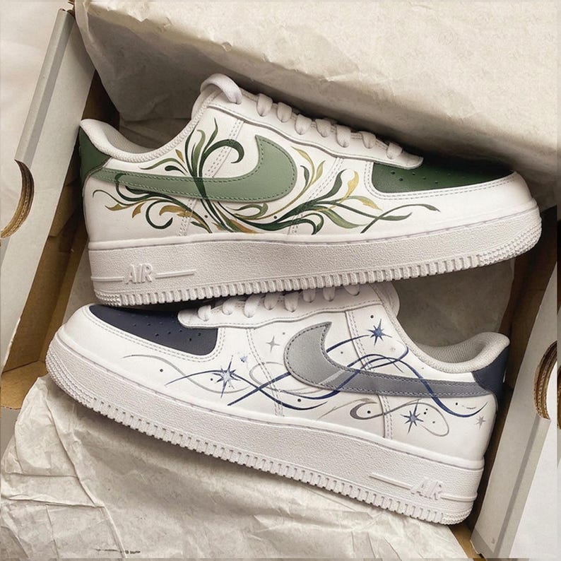 May include: White sneakers with custom painted designs. One pair features green and gold floral patterns, the other has a navy blue and gray celestial design with stars and swirls. Both pairs have white soles and the "AIR" logo.