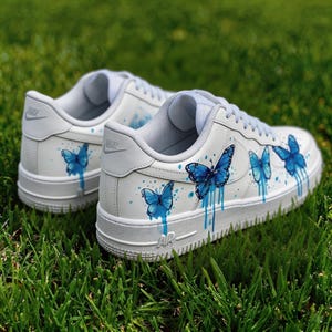 Op de afbeelding: Witte sneakers met blauwe vlinderontwerpen. De schoenen hebben een witte basis met lichtblauwe accenten op de tong en hiel. De vlinders zijn geschilderd in blauwtinten, met een druppelend effect.
