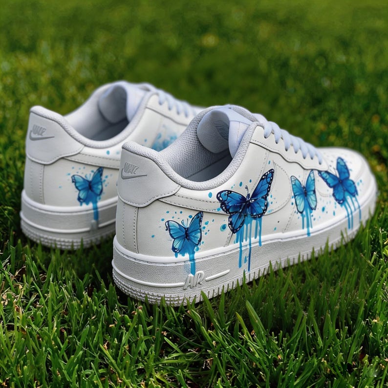 Op de afbeelding: Witte sneakers met blauwe vlinderontwerpen. De schoenen hebben een witte basis met lichtblauwe accenten op de tong en hiel. De vlinders zijn geschilderd in blauwtinten, met een druppelend effect.