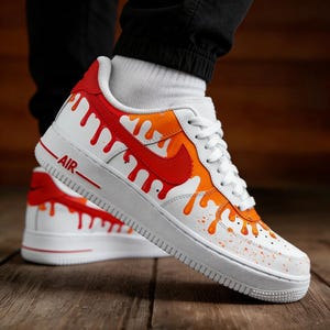 May include: White sneakers with red and orange paint drip design. The shoes have a red Nike swoosh and the word "AIR" in red. The soles are white, and the laces are white.