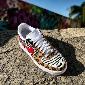 May include: A custom sneaker with a white base, zebra and leopard print patterns. It features a red swoosh logo and white laces. The sole is white. The shoe is displayed on a textured stone surface. The shoe's design is eye-catching.