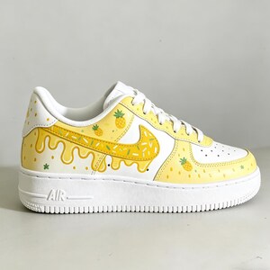 May include: White and yellow custom sneakers with a pineapple theme. The shoes feature a white base with yellow accents, including a dripping design and pineapple illustrations. The iconic Nike swoosh is also decorated with yellow and green details.