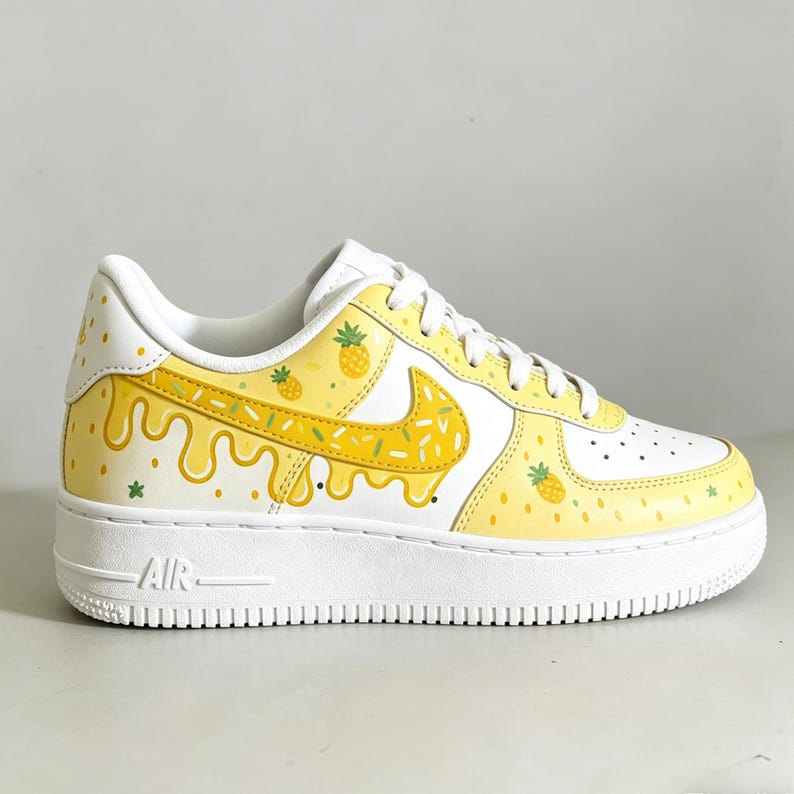 May include: White and yellow custom sneakers with a pineapple theme. The shoes feature a white base with yellow accents, including a dripping design and pineapple illustrations. The iconic Nike swoosh is also decorated with yellow and green details.