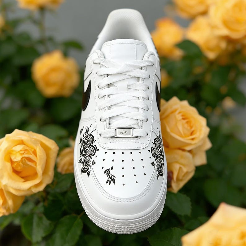 May include: White leather sneaker with black floral design and black swoosh. The shoe features a white sole, laces, and tongue. The sneaker is decorated with black roses and leaves. The background is filled with yellow roses.