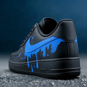 May include: Black and blue athletic shoe with a low-top design. The shoe features a black base with blue paint splatter details and a blue Nike swoosh logo on the side. The sole is black.