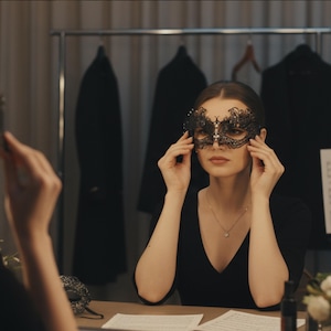 Op de afbeelding: Een zwart kanten masker met een bloemenontwerp en kristallen accenten. Het masker wordt door een persoon omhoog gehouden. Het masker ligt op een tafel met een spiegel die het masker weerspiegelt. De achtergrond omvat een kledingrek en een tafel.