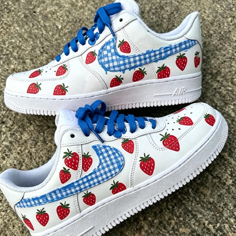 May include: White sneakers with red strawberry designs and blue laces. The Nike swoosh is a blue and white gingham pattern. The shoes have a white sole and the word "AIR" is visible on the side.