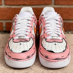 May include: Pair of pink and white sneakers with cartoon-style detailing. The shoes feature white laces, a white sole, and orange accents. The design includes black outlines and small perforations on the toe box.