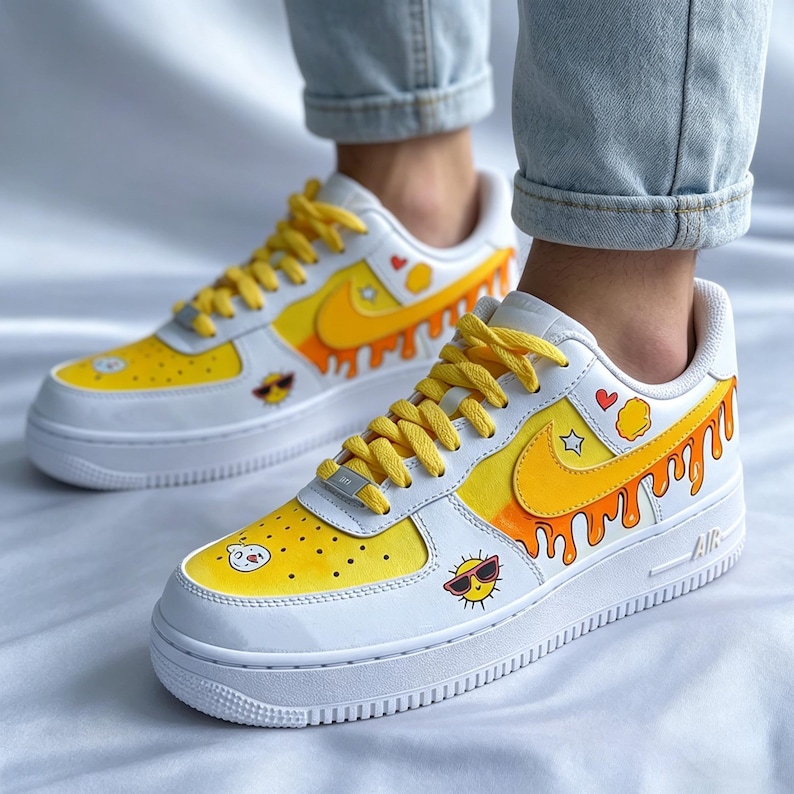 May include: White sneakers with yellow and orange painted accents. The design includes a yellow swoosh, dripping orange paint, and cartoon sun and heart graphics. The laces are yellow.