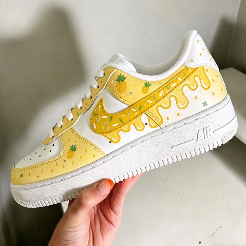 May include: White and yellow Air Force 1 sneakers with a pineapple theme. The sneakers feature a white base with yellow accents, including a dripping design, pineapple graphics, and a yellow Nike swoosh.