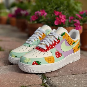 Puede incluir: Zapatillas blancas personalizadas con un dise&ntilde;o de frutas colorido. Los zapatos presentan fresas, pi&ntilde;as y otros patrones de frutas sobre una base blanca con un swoosh verde claro. Las suelas son blancas.