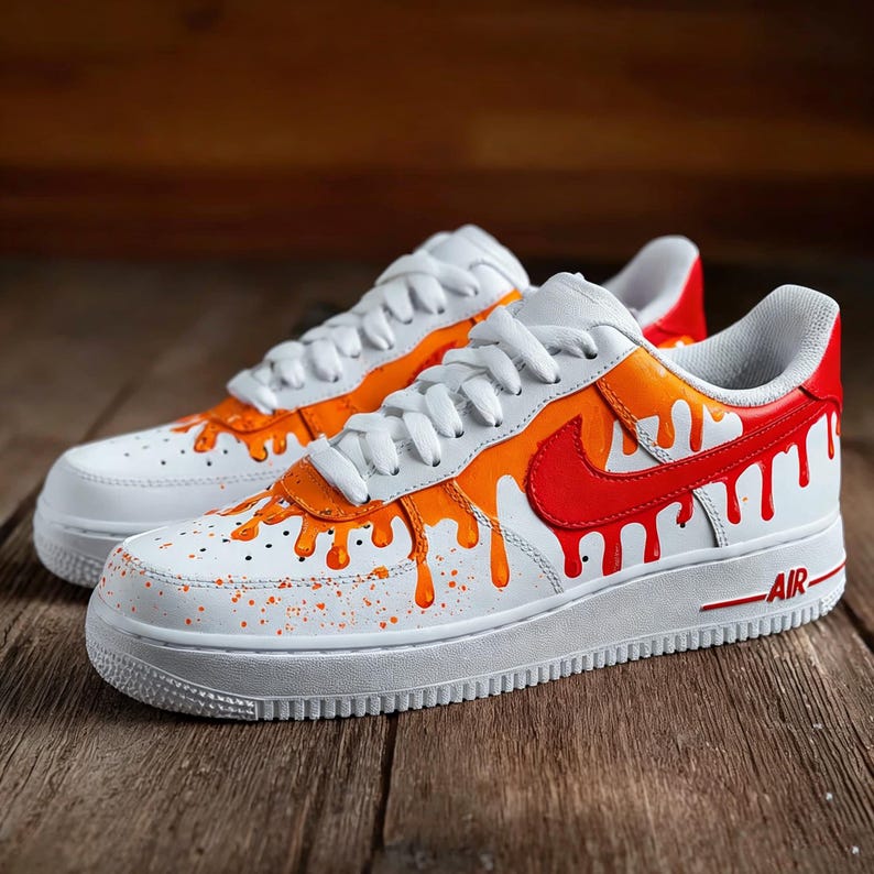 May include: White sneakers with orange and red paint drip design. The shoes have white laces, a white sole, and a red swoosh logo. The word "AIR" is printed in red on the side.