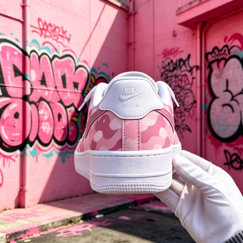 May include: Pink and white Nike Air Force 1 sneakers with a camouflage pattern. The sneakers have a white sole and a white Nike logo on the heel. The background features pink graffiti art.