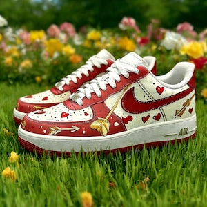 May include: Red and white sneakers with heart and arrow designs. The shoes feature a red swoosh, white laces, and a white sole with the word "AIR". The sneakers are on a bed of green grass with colorful flowers in the background.