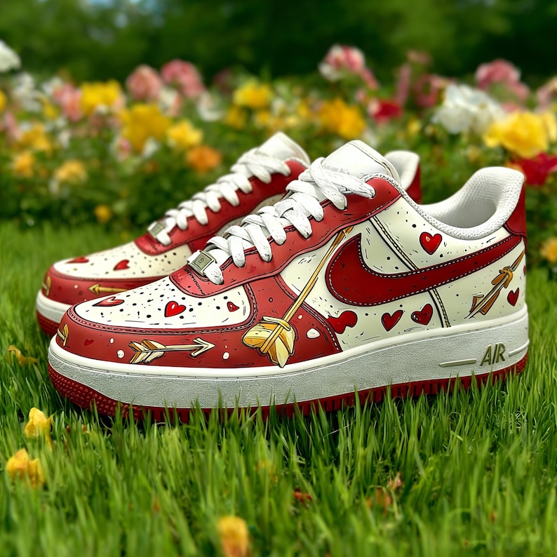 May include: Red and white sneakers with heart and arrow designs. The shoes feature a red swoosh, white laces, and a white sole with the word "AIR". The sneakers are on a bed of green grass with colorful flowers in the background.