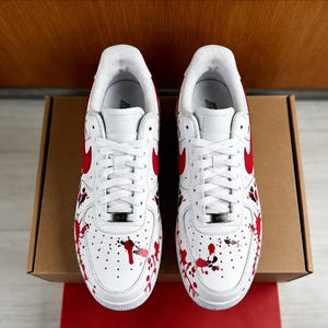 Op de afbeelding: Paar witte sneakers met rode accenten, inclusief de Nike swoosh en een verfspatterontwerp. De schoenen hebben witte veters en worden gepresenteerd op een kartonnen doos.