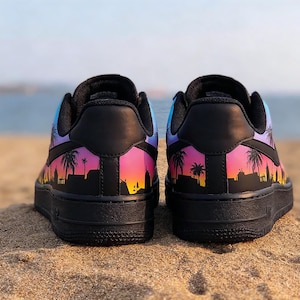 May include: Pair of black sneakers with a vibrant sunset design featuring palm trees and city silhouettes. The design transitions from purple to pink and orange, set against a blue sky. The shoes are on a sandy surface.