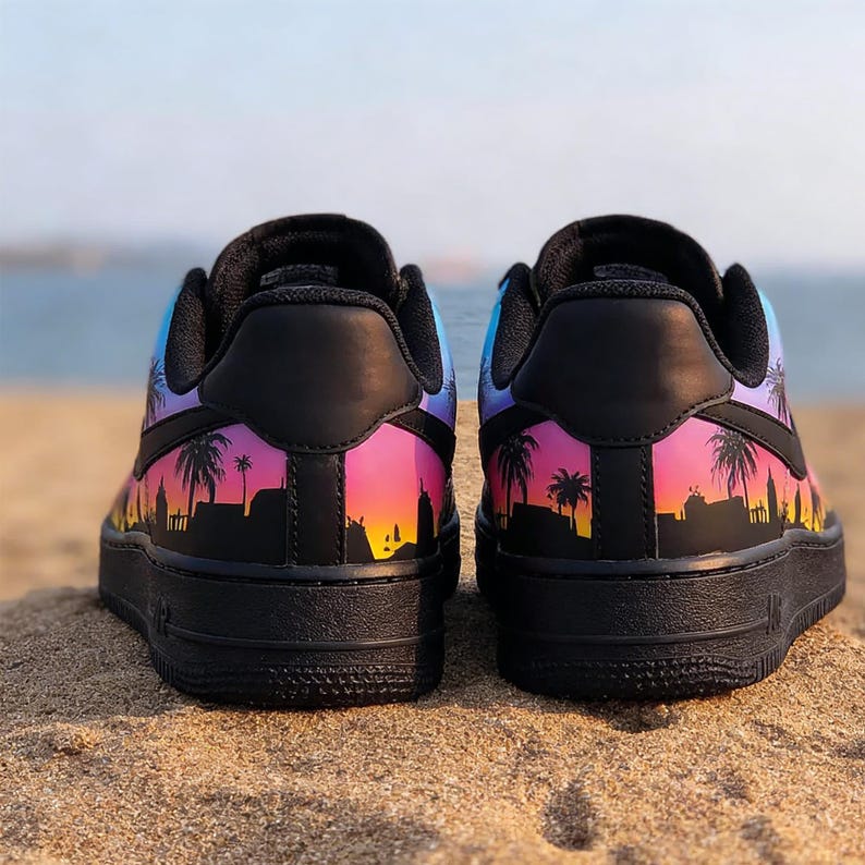 May include: Pair of black sneakers with a vibrant sunset design featuring palm trees and city silhouettes. The design transitions from purple to pink and orange, set against a blue sky. The shoes are on a sandy surface.