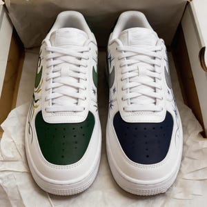 May include: Pair of white sneakers with custom designs. One shoe features a green toe and accents, the other a navy blue toe. Both have white laces and a silver swoosh. The sides are decorated with starburst and floral patterns.