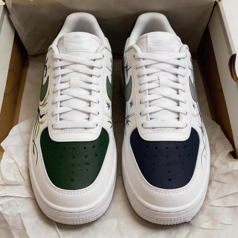 May include: Pair of white sneakers with custom designs. One shoe features a green toe and accents, the other a navy blue toe. Both have white laces and a silver swoosh. The sides are decorated with starburst and floral patterns.