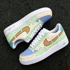 May include: Pair of custom Nike Air Force 1 sneakers with a unique ice cream cone design. The shoes feature a white sole, light blue, green, and yellow accents, and a brown waffle cone-shaped Nike swoosh with colorful sprinkles.