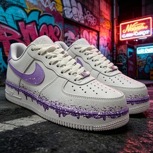 May include: A pair of white low-top sneakers featuring a lavender swoosh logo on the side, white laces, and a purple paint drip effect along the midsole. The shoes include a metallic lace dubrae and perforated toe boxes for a classic athletic look.