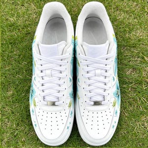 May include: Pair of white sneakers with blue floral designs. The shoes have white laces, a white sole, and a perforated toe box. The floral pattern includes blue flowers and green leaves, adding a touch of color to the classic design.
