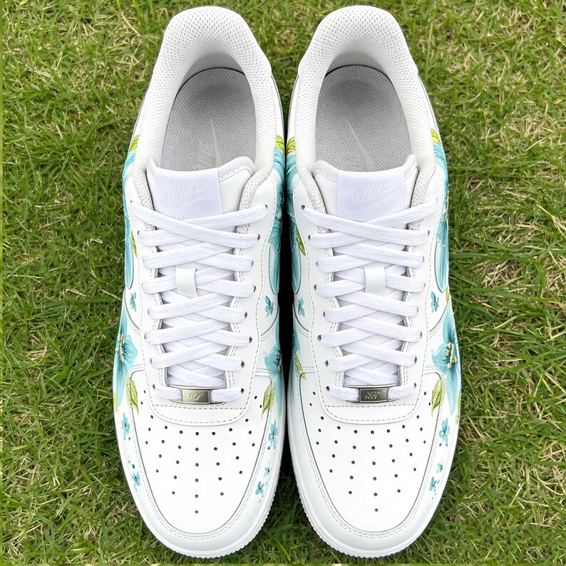 May include: Pair of white sneakers with blue floral designs. The shoes have white laces, a white sole, and a perforated toe box. The floral pattern includes blue flowers and green leaves, adding a touch of color to the classic design.