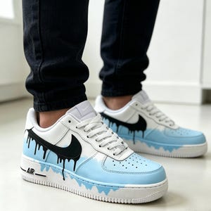 Puede incluir: Zapatillas blancas y celeste con detalles negros. Las zapatillas presentan un logotipo de Nike negro y un dise&ntilde;o &uacute;nico de pintura que gotea. La palabra "AIR" est&aacute; impresa en el lateral de la suela blanca.