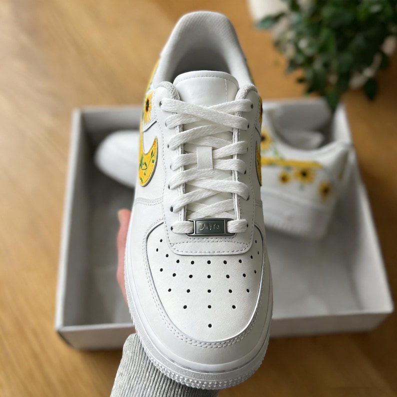 May include: White leather sneakers with yellow sunflower designs on the sides and heels. The sneakers have white laces, a white sole, and a small metal tag on the laces. The shoes are in a white box.