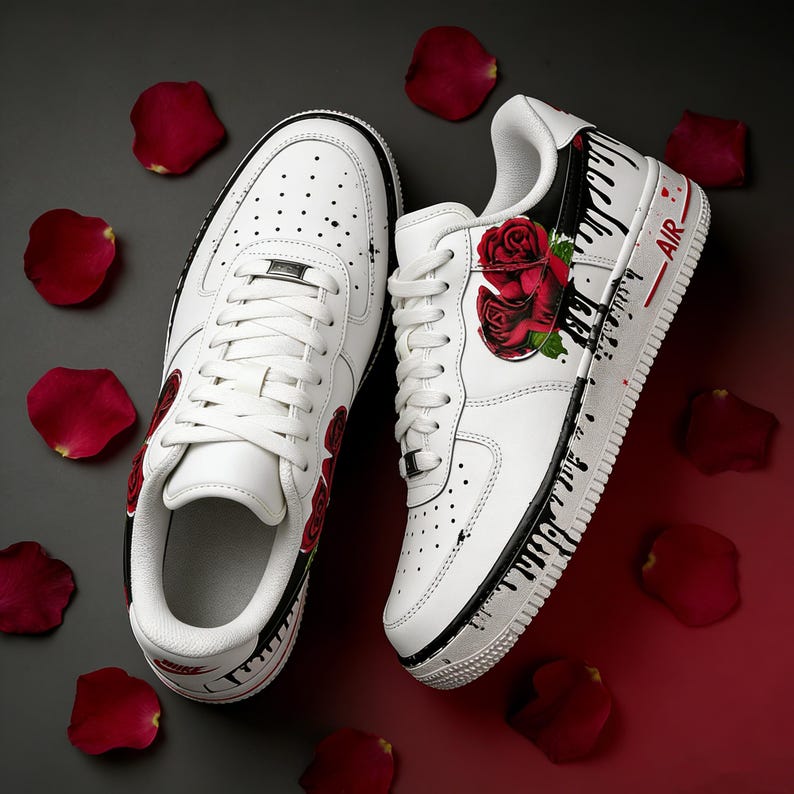 May include: White sneakers with red rose graphics and black paint drip details. The shoes have white laces, a white sole, and the word "AIR" in red. Red rose petals are scattered around the shoes.