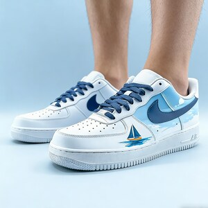 May include: White sneakers with blue accents, featuring a blue Nike swoosh and a nautical design of a sailboat on the side. The laces are blue, and the shoes are on a light blue background.