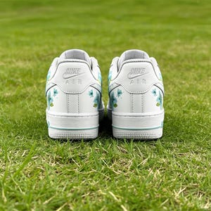 May include: A pair of white Nike Air Force 1 sneakers with floral accents. The shoes feature light blue and green floral designs on the sides and heels. The Nike logo is visible on the heel. The shoes are on a green grassy surface.