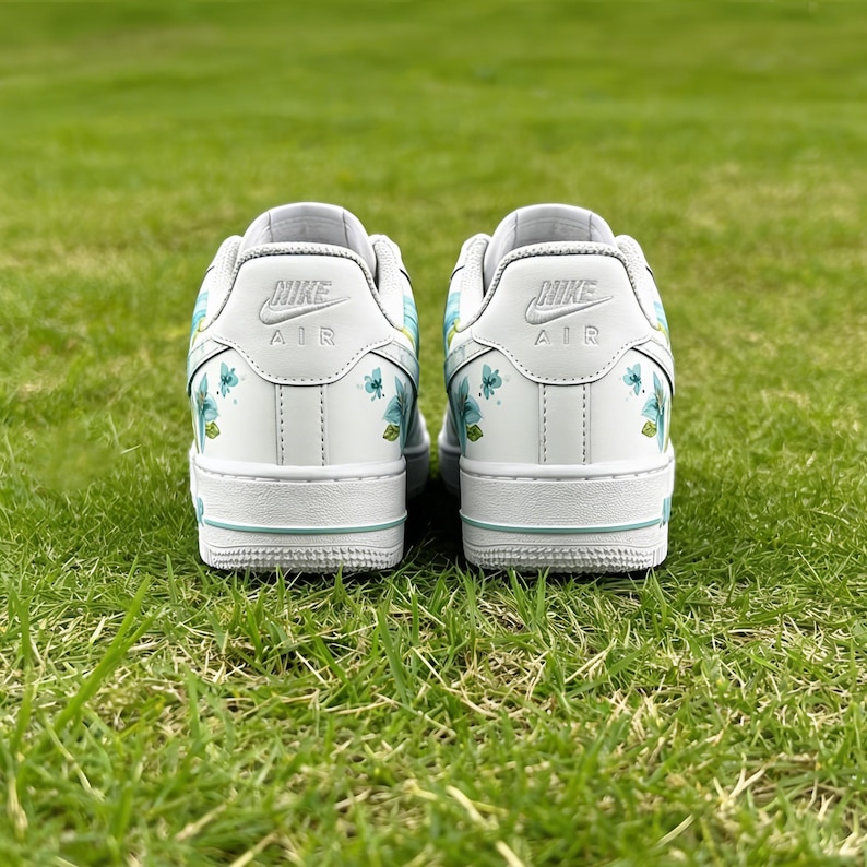 May include: A pair of white Nike Air Force 1 sneakers with floral accents. The shoes feature light blue and green floral designs on the sides and heels. The Nike logo is visible on the heel. The shoes are on a green grassy surface.