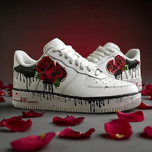 May include: White sneakers with a red rose design and black paint drip accents. The shoes have a classic low-top style with white laces and a red "AIR" detail on the midsole. Red rose petals are scattered around the shoes.