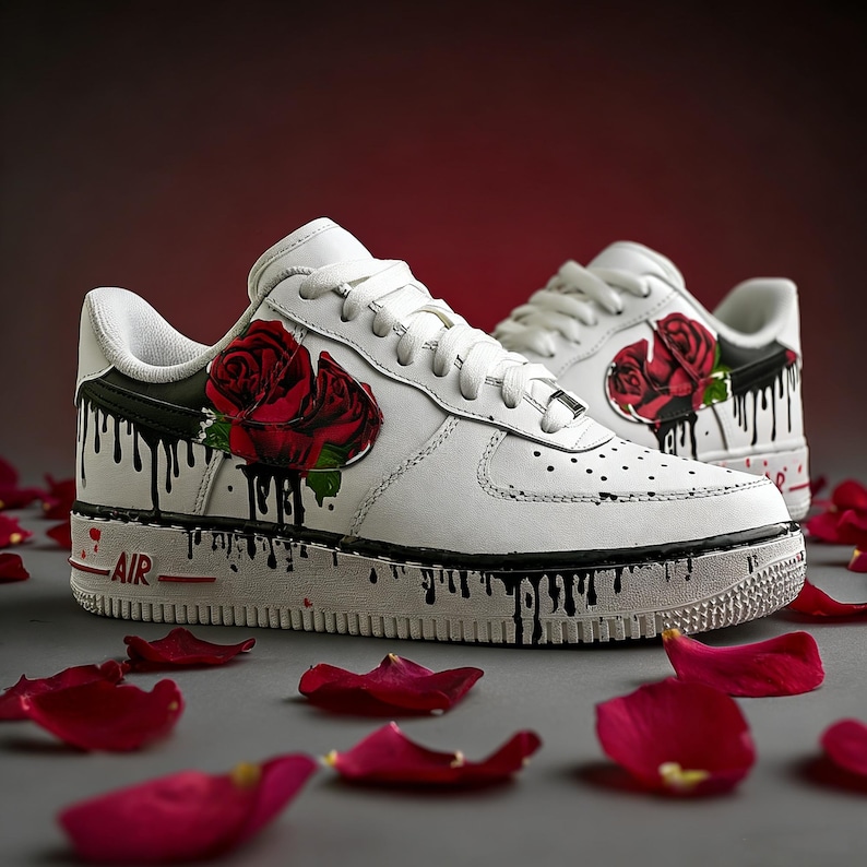 May include: White sneakers with a red rose design and black paint drip accents. The shoes have a classic low-top style with white laces and a red "AIR" detail on the midsole. Red rose petals are scattered around the shoes.