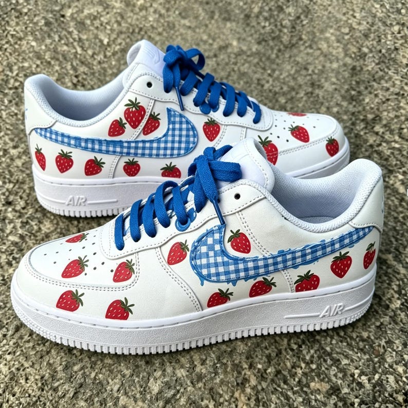 May include: White sneakers with blue laces and a strawberry pattern. The shoes feature a blue and white gingham check design on the side and red strawberry graphics. The soles are white.