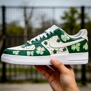 May include: White and green athletic shoe with a clover design. The shoe features a white sole, green accents, and a cream-colored Nike swoosh. The upper is white with green shamrock graphics. The laces are white.
