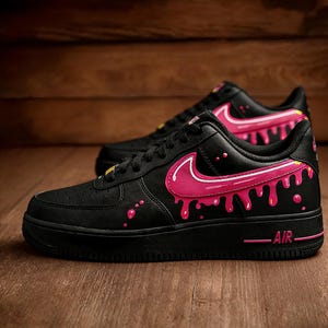May include: Pair of black sneakers with pink dripping paint design. The shoes feature a pink Nike swoosh and pink paint drips along the sides. The word "AIR" is printed in pink on the sole.
