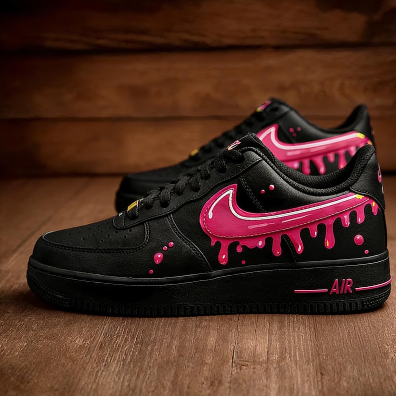 May include: Pair of black sneakers with pink dripping paint design. The shoes feature a pink Nike swoosh and pink paint drips along the sides. The word "AIR" is printed in pink on the sole.