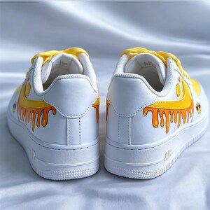 May include: Pair of white sneakers with yellow laces and a yellow and orange dripping design. The shoes have a white sole and the word "AIR" is visible on the side. The design resembles honey.
