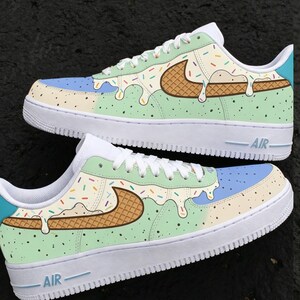 May include: Custom-painted sneakers with an ice cream theme. The shoes feature a white base with a light blue and cream-colored toe box. The sides are mint green with a waffle cone design and sprinkles, and a melting ice cream effect.