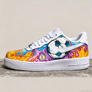 May include: White athletic shoe with a colorful, custom design. The shoe features a soccer ball graphic, flames, and a compass. The design incorporates shades of blue, yellow, orange, and purple. The shoe has white laces and a white sole.