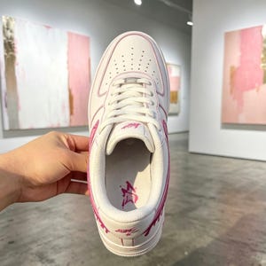 May include: White and pink leather sneaker with a low-top design. The shoe features pink accents on the stitching, logo, and sole. The interior has a pink logo design. The shoe is displayed in a gallery setting.