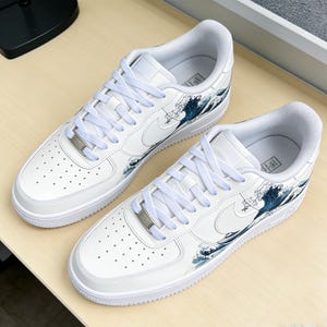 May include: Pair of white sneakers with a wave design in shades of blue. The shoes feature white laces, a silver metal tag, and the Nike swoosh logo. The design is inspired by the famous Japanese artwork, "The Great Wave off Kanagawa."