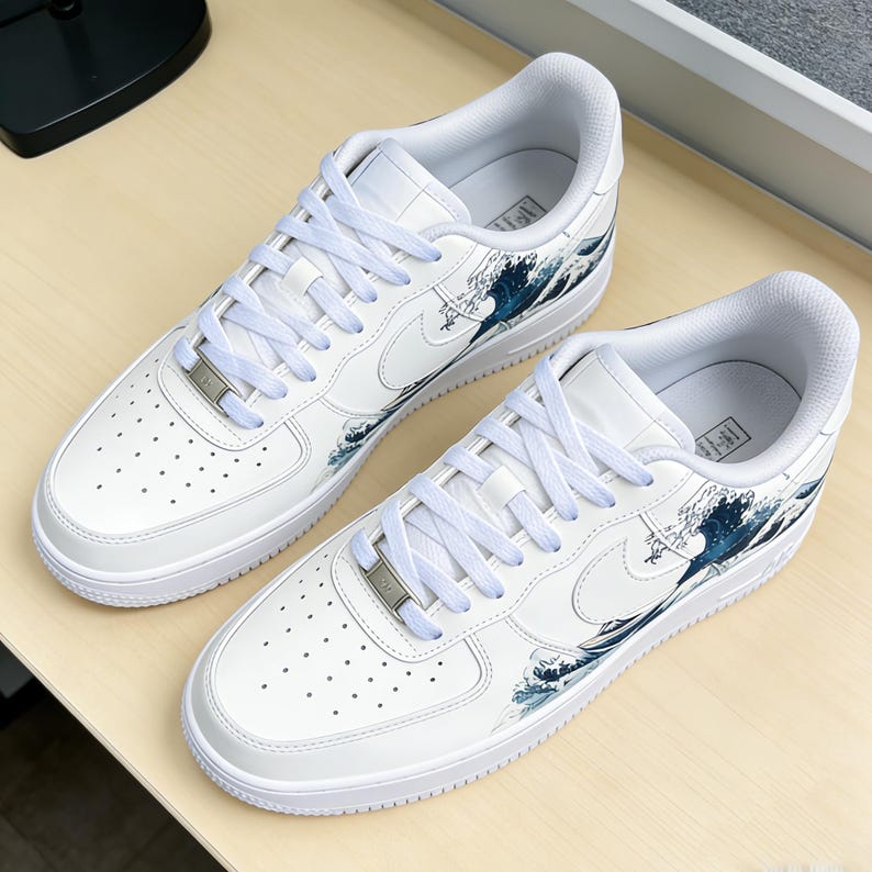 May include: Pair of white sneakers with a wave design in shades of blue. The shoes feature white laces, a silver metal tag, and the Nike swoosh logo. The design is inspired by the famous Japanese artwork, "The Great Wave off Kanagawa."