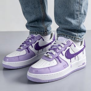 May include: White and purple sneakers with a drip design. The sneakers feature a white base with purple accents, including the Nike swoosh and a dripping paint effect. A small heart is visible on the side of the shoe.